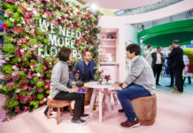 An image of IPM Essen 2020, three persons sitting in a table at a flower wall with the description in it: We need more flowers