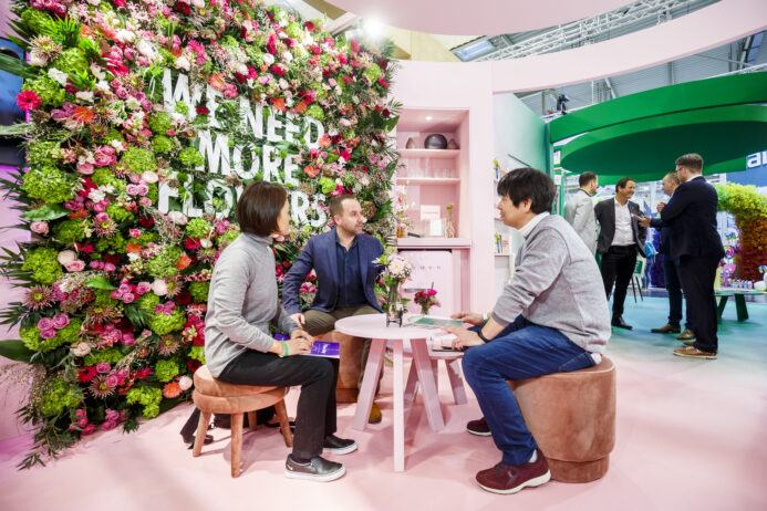 An image of IPM Essen 2020, three persons sitting in a table at a flower wall with the description in it: We need more flowers